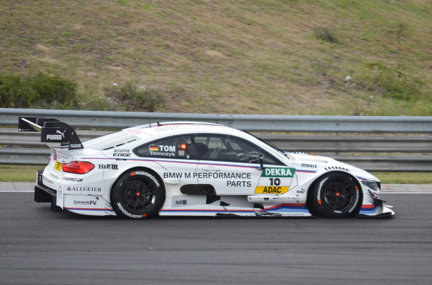 Ein BMW M4 DTM Rennauto auf einer Rennstrecke mit Text auf der Karosserie, umgeben von grünem Gras und einer Begrenzung im Hintergrund.