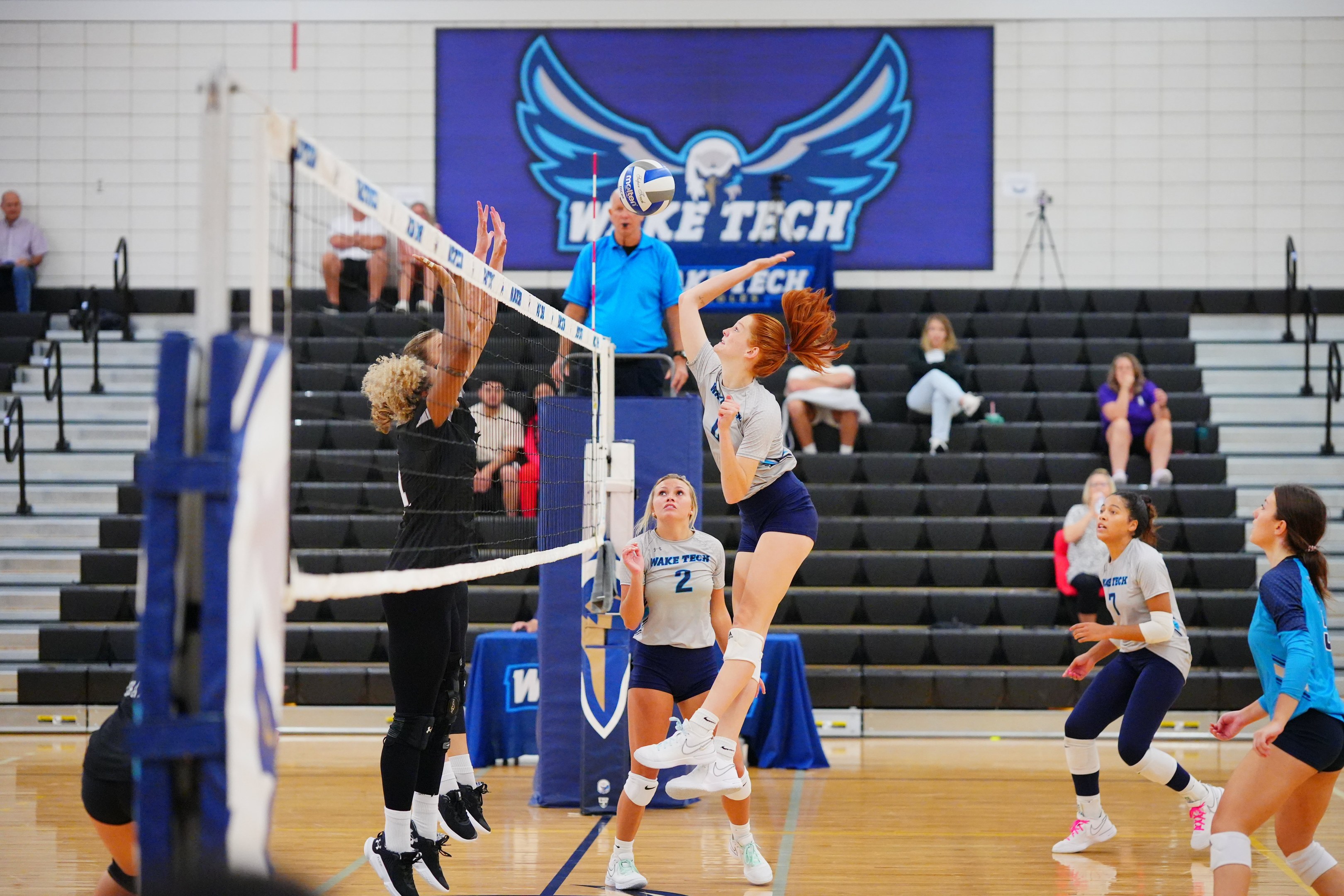 Frauen spielen Volleyball mit einem Netz, Zuschauer auf Treppe und ein Meisterschaftsbanner mit einem Adler an der Wand.