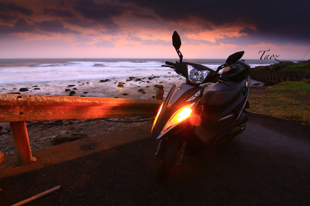 Ein Roller steht auf der Seite eines Straßenrandes neben dem Ozean, neben einem Zaun, Gras, Steinen und Wasser im Hintergrund, unter einem bewölkten Himmel, mit einem Wasserzeichen.