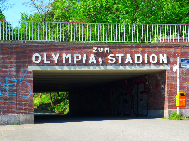 Der Eingang zum Olympiastadion in Berlin, Deutschland, mit einer Brücke mit Text, einem Metallzaun, einer Schautafel, einer Box, Pflanzen, Gras, Bäumen und einem bewölkten Himmel.