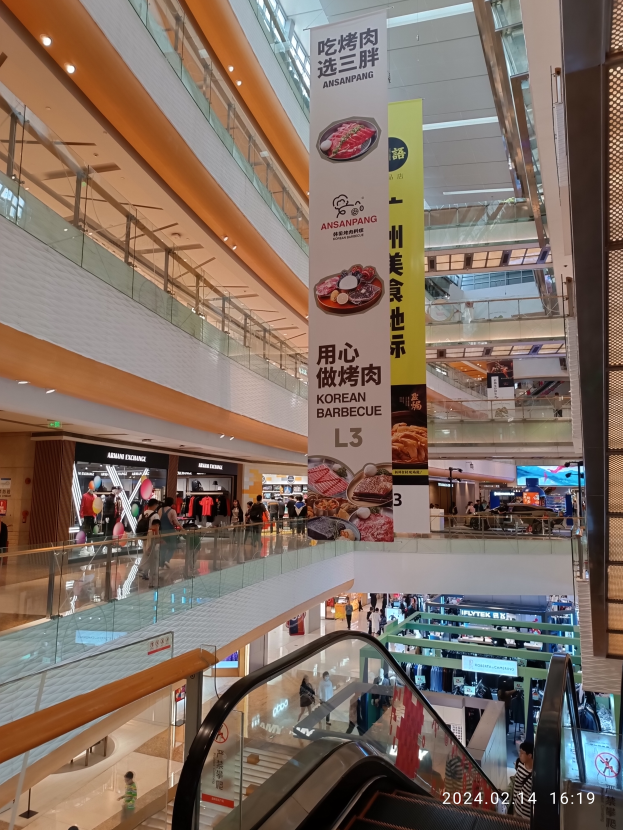 Einblicke in ein Einkaufszentrum mit einer Rolltreppe in der Mitte, Menschen, die herumlaufen, einige mit Handtaschen, und Schilder von der Decke hängen, mit Geschäften im Hintergrund.