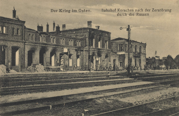 Altes Schwarz-Weiß-Foto des Bahnhofs Korschen in Ostern, Deutschland, zeigt einen Bahnhof mit Gleisen, Gebäuden, Masten, Menschen, Bäumen und Himmel im Hintergrund.
