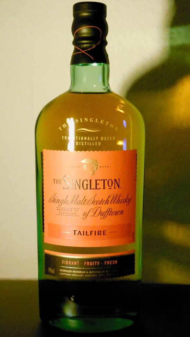 Eine tiefe Bernsteinflasche Singleton of Dufftown Tailfire Single Malt Scotch Whisky auf einem Tisch, mit einem schwarzen Etikett mit fetter Schrift und einem Wappen mit einem Löwen und einem Schloss.