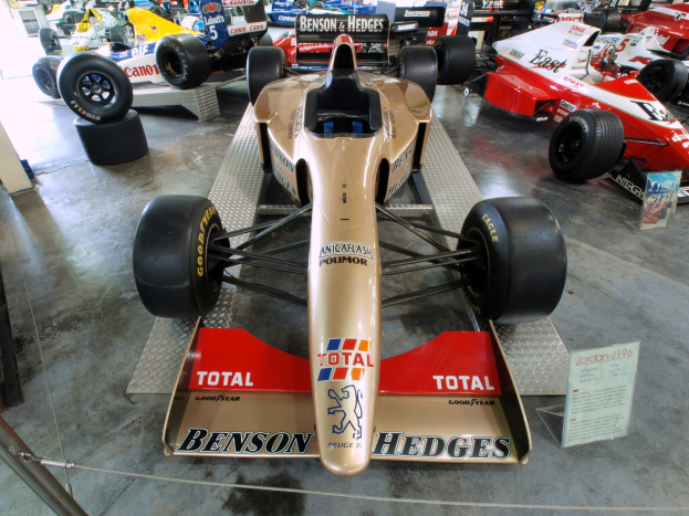 Rennwagen in einem Motorsportmuseum ausgestellt, getrennt durch einen Metallzaun mit Informationsschild im Vordergrund und Besuchern im Hintergrund.