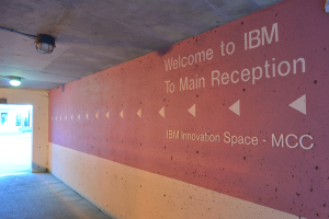 Eine Wand mit 'Willkommen bei IBM' beleuchtet von Deckenlampen, mit einer Tür zum IBM Innovation Space MCC im Hintergrund.