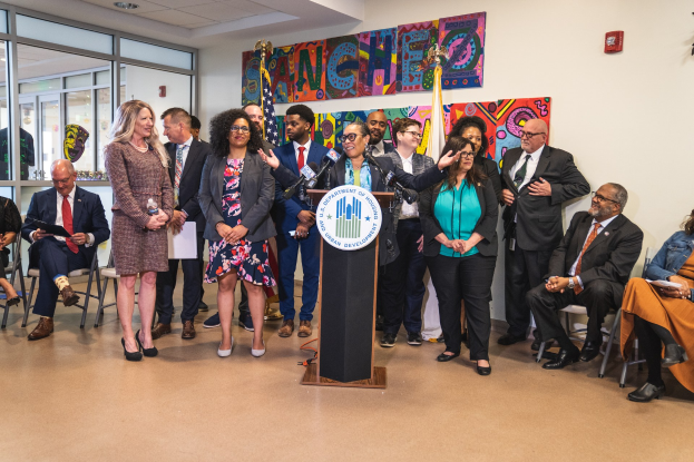 Gruppe von Menschen vor einem Podium mit einem Mikrofon und einer Tafel mit der Aufschrift "Sancho City Councilman Joins City Council", mit sitzenden und stehenden Zuschauern, Flaggen, einem Feuerlöscher und Deckenleuchten im Hintergrund.