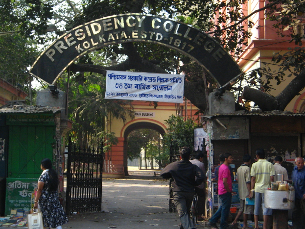 Der Eingang zum Presidency College in Kolkata, Indien, mit einem Bogen mit einem 'Presidency College'-Banner, einem Tor, Menschen, die auf der rechten Seite gehen, und einem Gebäude mit Bäumen unter einem klaren blauen Himmel im Hintergrund.