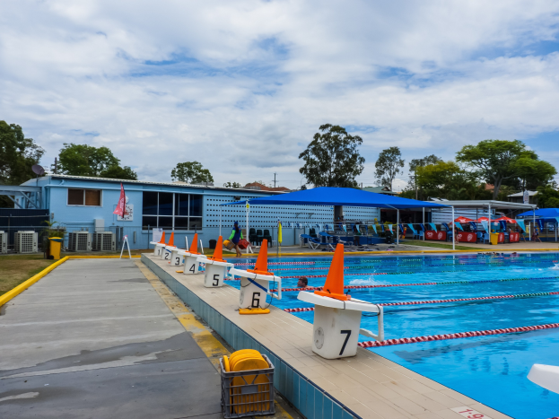 Großer Swimmingpool mit mehreren Schwimmern, Bahnteiler, Verkehrskegeln, Liegestühlen, Sonnenschirmen, einem Gebäude mit Fenstern, einer Flagge, Bäumen und einem bewölkten Himmel im Hintergrund.