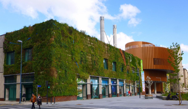 Ein großes Gebäude mit einer grünen Wand an seiner Seite, umgeben von Straßenmasten, Straßenlaternen, Verkehrspfosten, Verkehrsampeln, Kraftfahrzeugen, Fußgängern, Bäumen und einem bewölkten Himmel, als Teil des Green Building Project.