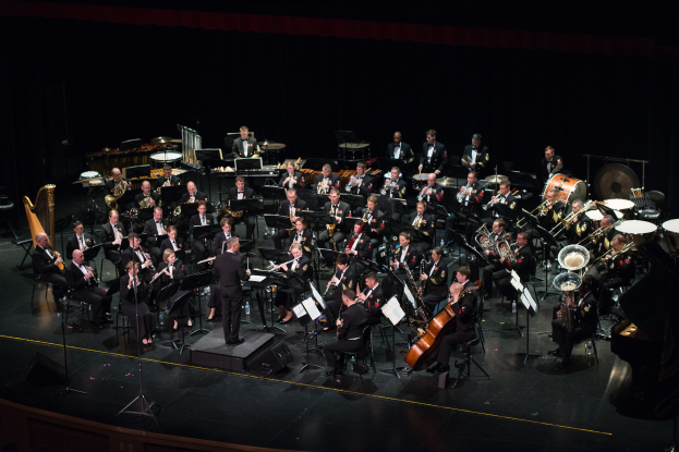 Ein großes Orchester auf einer Bühne mit einem Dirigenten in der Mitte, mit verschiedenen Instrumenten wie Saxophone, Trompeten, Posaunen und Trommeln, und der Dirigent in einem schwarzen Anzug und weißem Hemd unter hellen Bühnenlichtern und einem Publikum im Hintergrund.