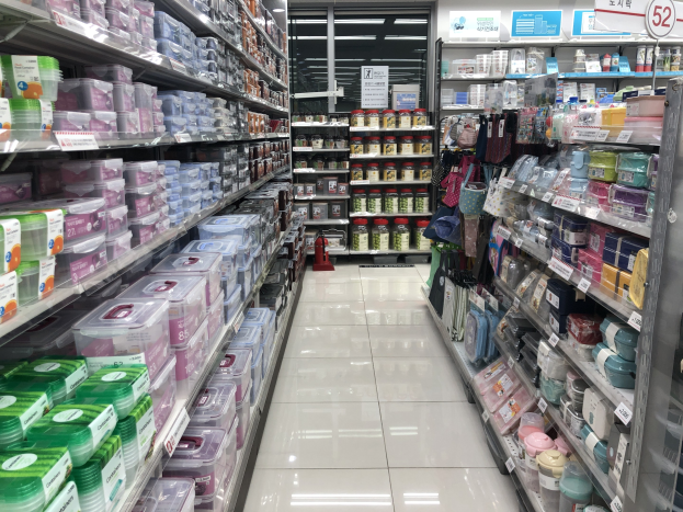 Ein gut sortierter chinesischer Supermarktgang mit Regalen voller Schachteln, Flaschen, Taschen und anderen Produkten, beleuchtet von Deckenlampen und gekennzeichnet durch Texttafeln, einschließlich eines sichtbaren Feuerlöschers auf dem Boden.