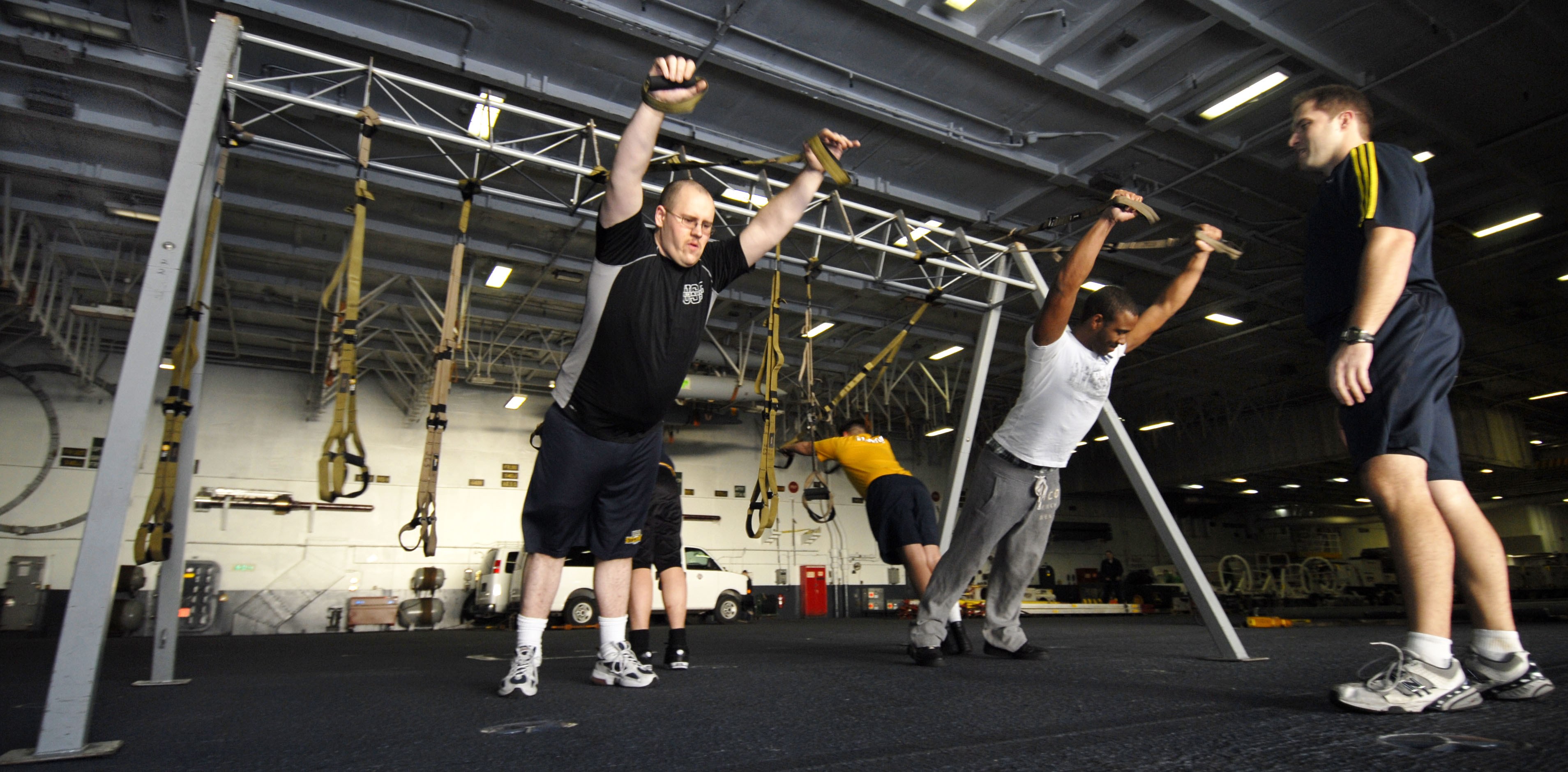 Gruppe von Männern, die an Stangen in einem Fitnessstudio mit Deckenbeleuchtung und verschiedenen Gegenständen im Hintergrund Pull-ups machen, wobei ein Mann in der Mitte Schuhe trägt.