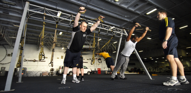 Gruppe von Männern, die an Stangen in einem Fitnessstudio mit Deckenbeleuchtung und verschiedenen Gegenständen im Hintergrund Pull-ups machen, wobei ein Mann in der Mitte Schuhe trägt.
