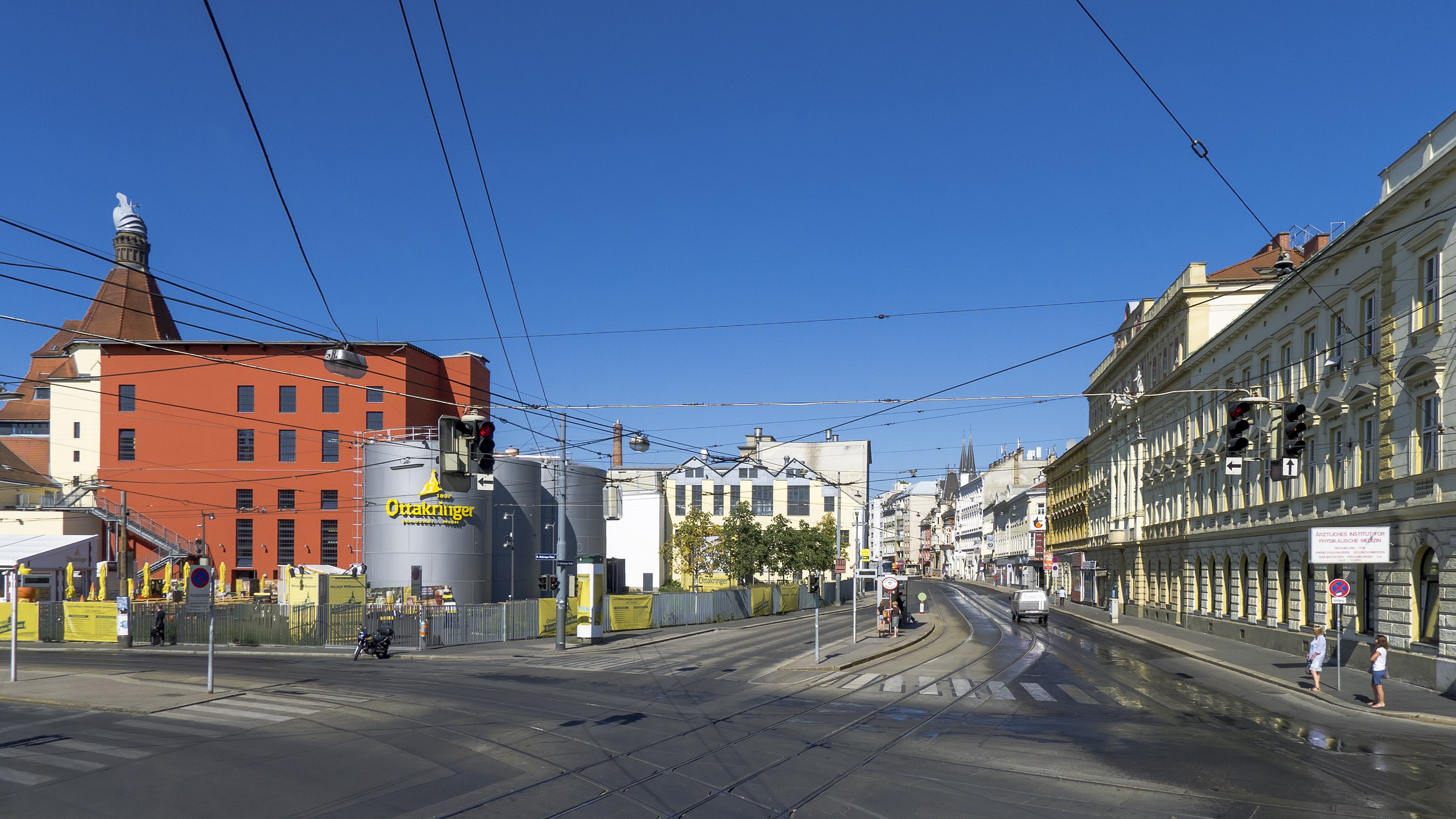 Eine Straße in der Stadt mit Gebäuden, Laternenmästen, Lichtern, Ampeln, Strommästen, Kabeln, fahrenden Fahrzeugen, Passanten auf dem Gehweg, Bäumen und Himmel im Hintergrund.