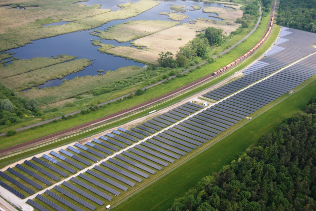 Luftaufnahme einer Solaränlage mit Panelen, umgeben von Bäumen, Gras, Pflanzen und Wasser, mit einem Zug auf einer näheren Eisenbahnschiene.