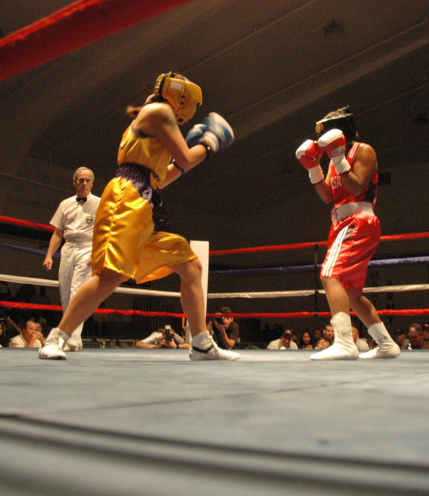 Zwei Boxer kämpfen in einem Ring mit einem Schiedsrichter und Zuschauern, einige halten Kameras, unter einer sichtbaren Decke.