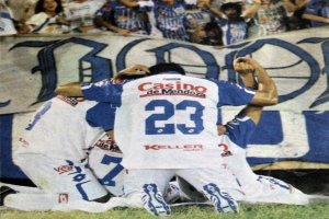 Gruppe von Füßtballspielern, die auf dem Boden liegen und mit jubelnden Fans im Hintergrund feiern, im Hintergrund ist ein Banner mit Text zu sehen.