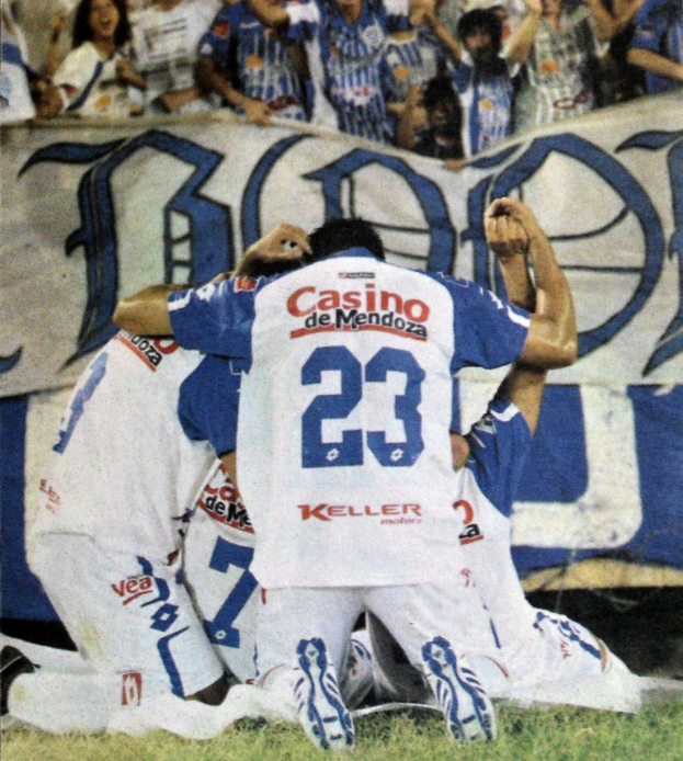 Gruppe von Füßtballspielern, die auf dem Boden liegen und mit jubelnden Fans im Hintergrund feiern, im Hintergrund ist ein Banner mit Text zu sehen.