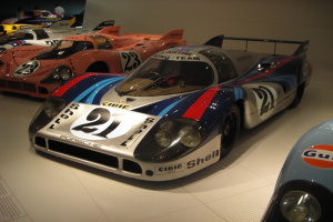 Ein Porsche 917K Le Mans Rennwagen wird in einem Museum ausgestellt, umgeben von anderen Sportwagen und Menschen, mit einer Wand im Hintergrund und einem Blatt Papier mit Text auf der linken Seite.