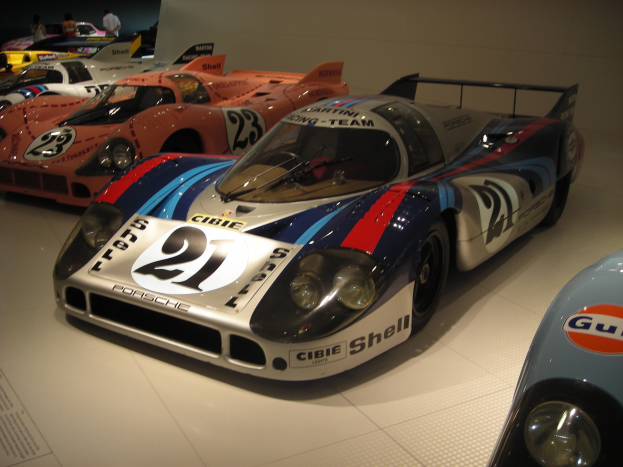 Ein Porsche 917K Le Mans Rennwagen wird in einem Museum ausgestellt, umgeben von anderen Sportwagen und Menschen, mit einer Wand im Hintergrund und einem Blatt Papier mit Text auf der linken Seite.
