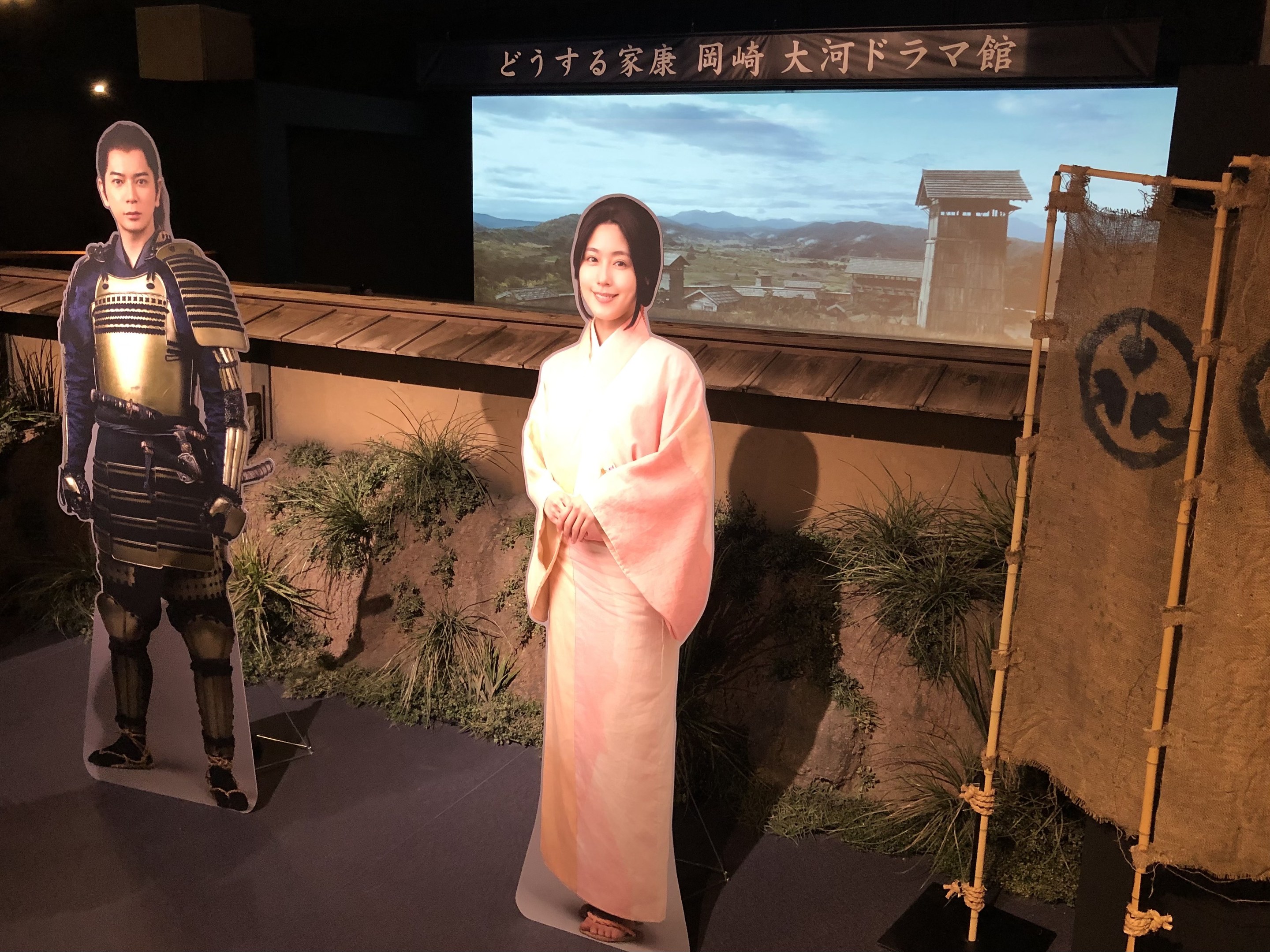 Zwei Menschen – eine Frau in einem rosa Kimono und ein Mann in Samurai-Rüstung – lächeln gemeinsam in einer Szene aus einem japanischen Film, mit Pflanzen, einem Bildschirm und Beleuchtung im Hintergrund.