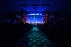 Ein großer Auditorium mit Sitzplätzen, eine Bühne mit Musikern und eine Decke mit Lichtern und Fernsehern, was auf einen Konzertsaal für die Nationalkonvention 2019 hindeutet.
