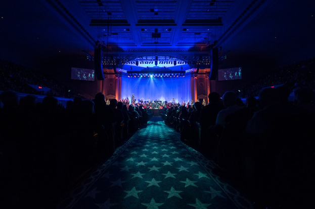 Ein großer Auditorium mit Sitzplätzen, eine Bühne mit Musikern und eine Decke mit Lichtern und Fernsehern, was auf einen Konzertsaal für die Nationalkonvention 2019 hindeutet.