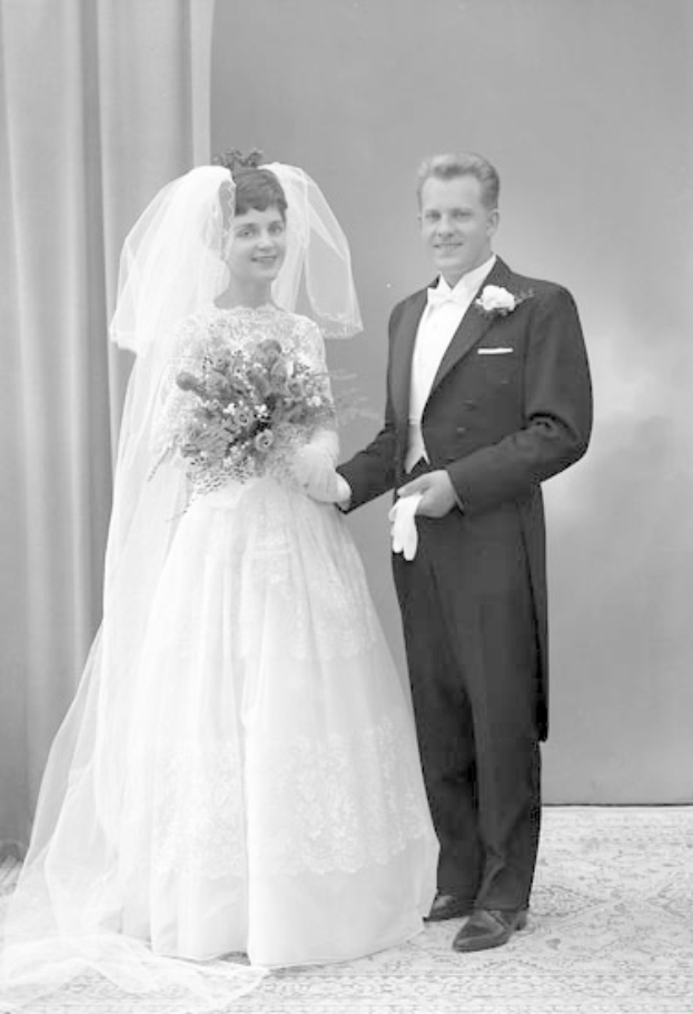 Ein Schwarz-Weiß-Foto eines lachenden Mannes und einer Frau, die an ihrem Hochzeitstag nebeneinander stehen, wobei die Frau ein weißes Kleid trägt und einen Strauß hält und eine Vorhangwand im Hintergrund zu sehen ist.