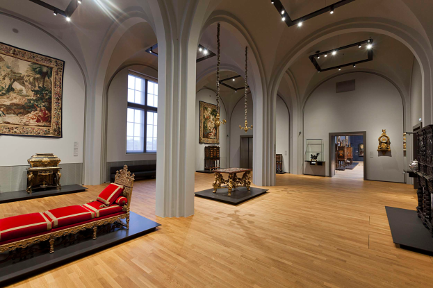 Ein geräumiger Raum im Kunsthistorischen Museum in Wien, Österreich, mit einer Couch mit Kissen, Tischen mit verschiedenen Gegenständen, Säulen, Fenstern und Deckenleuchten sowie zahlreichen paintings an den Wänden.