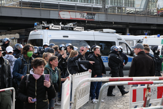 Eine Gruppe von Menschen steht in der Nähe von Polizeiwagen und Barrieren, einige tragen Helme und halten Telefone, mit einer Brücke und Gebäuden im Hintergrund während einer Demonstration in Berlin.