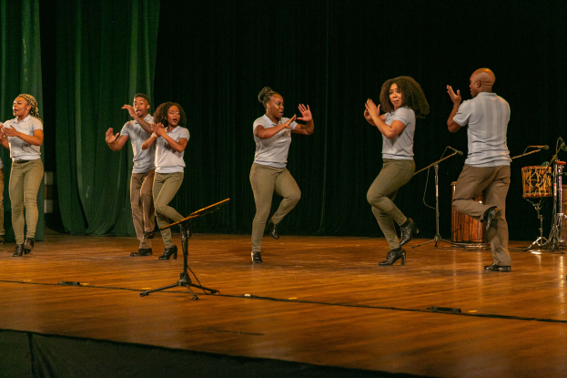 Eine Gruppe von Menschen auf einer Bühne mit Mikrofonen, Musikinstrumenten und einem Vorhang im Hintergrund, die Hände klappend in Feierlaune, scheinbar ein Tanzteam.