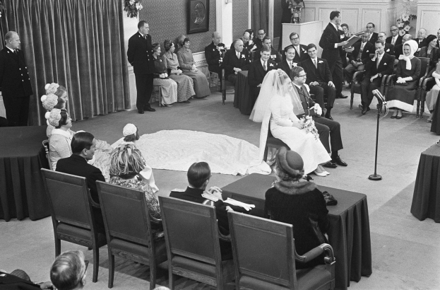 Schwarzes und weißes Foto einer Hochzeitszeremonie mit sitzenden und stehenden Menschen, einem Mann mit Mikrofon und einer Frau mit Blumenstrauß, mit einem Rednerpult, Blumenschmuck, einem Vorhang, einem gerahmten Foto an der Wand und einer Tür im Hintergrund.