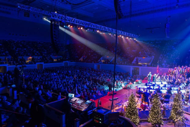 Eine große Menschenmenge in einem geschmückten Auditorium schaut einer Band zu, die auf einer Bühne mit Weihnachtsbäumen, Lichtern und Lautsprechern spielt.