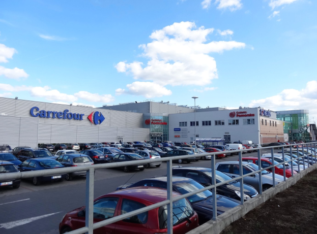 Großer Autohandel mit mehreren geparkten Fahrzeugen vor einem städtischen Hintergrund, umgeben von Straßenlaternen, Straßenschildern und Fahrzeugen auf der Straße unter einem bewölkten Himmel.