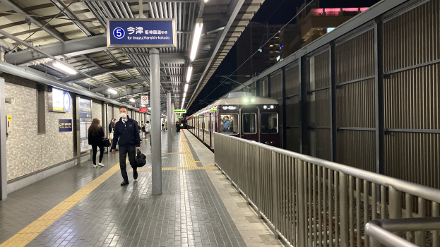 Ein Zug fährt auf einem Bahnsteig ein, auf dem Menschen gehen, ein Geländer auf der rechten Seite, Gebäude im Hintergrund und Leuchten mit einer Texttafel oben auf dem Bahnsteig.