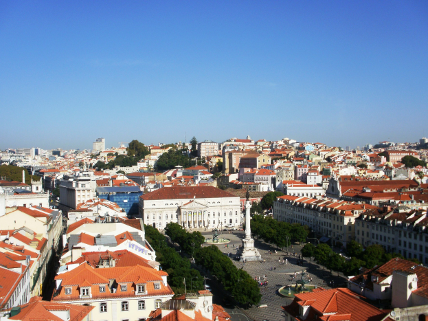 Blick auf Lissabon von einem Hügel mit Gebäuden mit Fenstern, Bäumen, einer Statue auf einem Sockel, Menschen auf der Straße und dem Himmel im Hintergrund.