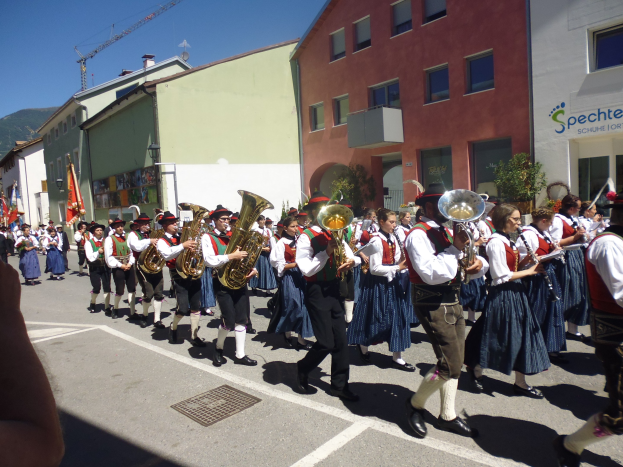 Eine Gruppe von Menschen in traditioneller bayrischer Tracht, die auf einer von Gebäuden gesäumten Straße Musikinstrumente spielen und dabei einen Hügel und einen klaren blauen Himmel im Hintergrund haben.