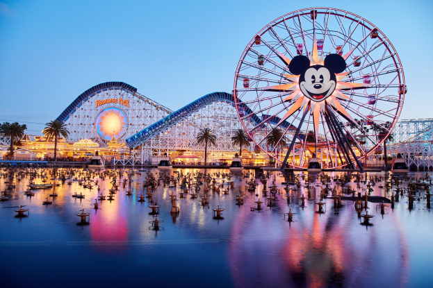 Eine farbenfrohe Dämmerungsszene im Disneyland California Adventure mit einem riesigen Mickey-Maus-Rad im Vordergrund, einer Achterbahn im Hintergrund und Booten auf dem Wasser, mit einem bunten Himmel und silhouettenartigen Bäumen.