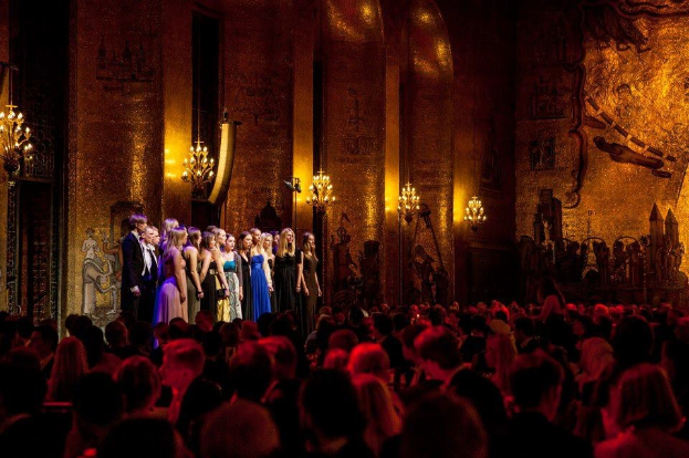Eine Gruppe von Menschen auf einer Bühne im Royal Opera House in London, mit beleuchteten Lichtern und Wandmalereien, die auf ein sitzendes Publikum schauen.