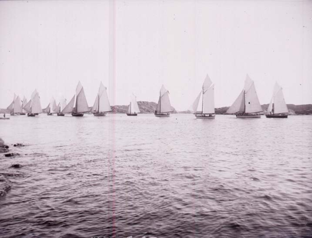 Schwarzes und weißes Foto von Segelbooten, die auf dem Wasser bei einem klaren Himmel und Bergen im Hintergrund rasen, beschriftet als New York Yacht Club.