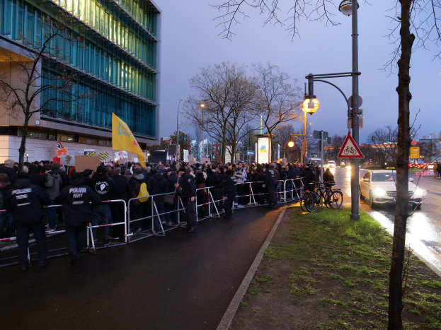 Große Gruppe von Menschen protestiert vor einem Gebäude in Berlin, hält Schilder hoch, mit Barrikaden, Fahrrädern, Laternen, Schildern, Bäumen und Gras im Hintergrund.