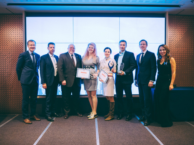 Eine Gruppe strahlender Menschen mit Urkunden, die vor einem Bildschirm stehen, mit einem Tisch, der mit schwarzem Tuch bedeckt ist und einem Wandobjekt im Hintergrund.