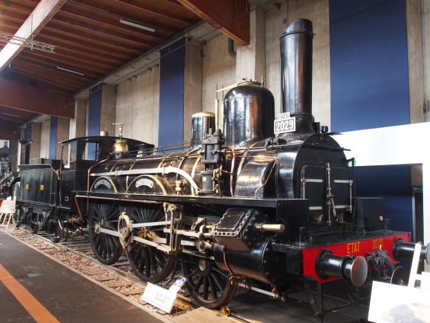 Alte Dampflokomotive in einem Museums Setting mit umgebenen Eisenbahnschienen, Informationsschildern und anderen Ausstellungsstücken an einer Wand mit Deckenbeleuchtung.