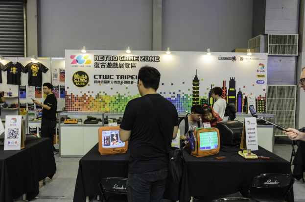 Gruppe von Menschen um einen Tisch versammelt, der ein Videospiel spielt, mit Stühlen, einer Wand, aufgehängten T-Shirts und Deckenbeleuchtung.