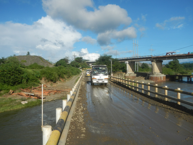 Ein Lkw fährt durch eine überflutete Straße neben einer Brücke mit Geländern, mit Bäumen, Strommasten und einem bewölkten Himmel im Hintergrund.