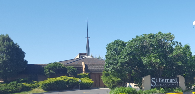 St. Bernard Catholic Church in San Antonio, Texas, ein Gebäude mit einem Kreuz darauf, umgeben von Pflanzen, Gras, Bäumen, einem Straßenpfahl, einer Schautafel, Straßenlampen und einem bewölktem Himmel.
