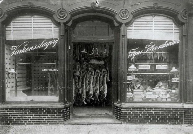 Schwarzes und weißes Foto einer Ladenfront mit einer Tür, Fenstern und einem Schankbrett, das ein belebtes Marktinnere mit verschiedenen Gegenständen zeigt.