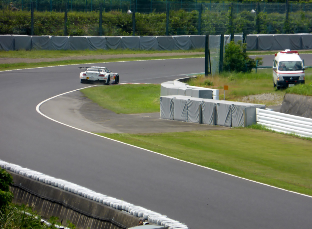 Ein Rennwagen auf einer Rennstrecke mit einem Polizeiwagen im Hintergrund, umgeben von Gras, Pflanzen, Bäumen und Zäunung, mit einem Fahrzeug auf der rechten Seite und Text unten.