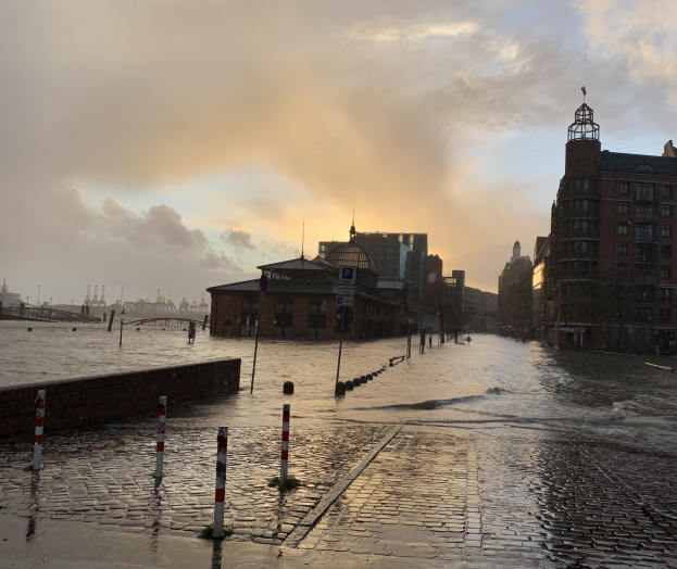 Überschwemmte Straße in Hamburg, Deutschland, mit Wasser, das die Straße, Pfähle, Schilder, Gebäude mit Fenstern, eine Brücke und einen bewölkten Himmel bedeckt.