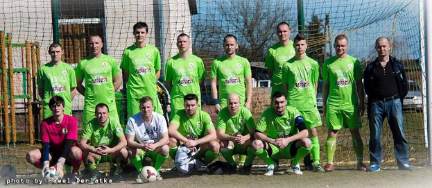 Eine Gruppe von Männern in grünen T-Shirts steht und hockt auf einem Fußballfeld mit einem Ball vor ihnen, ein Torpfosten rechts und ein Hintergrund aus Bäumen, Gebäuden, Pfählen, Fahrzeugen und dem Himmel, mit einer Zahlenfolge links unten.
